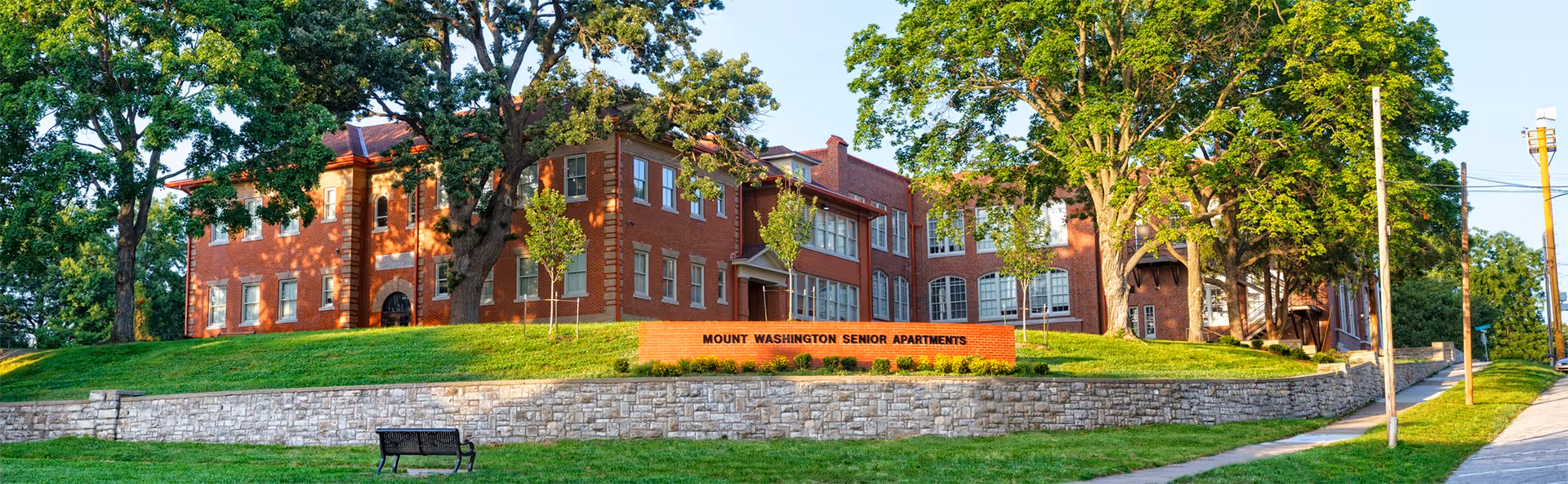 Brick senior apartments building on a grassy slope with a stone wall, trees, and bench.