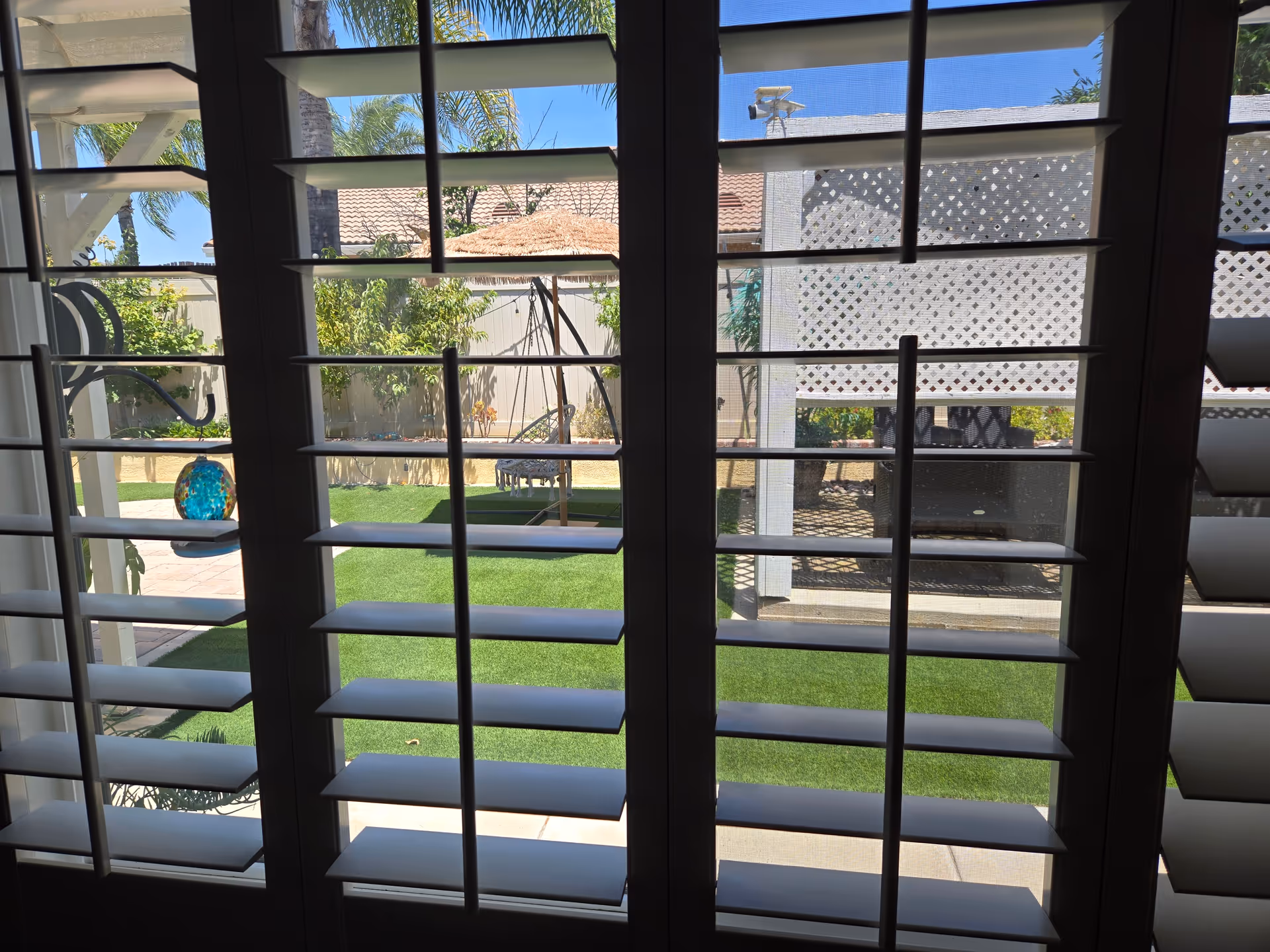 View from inside through louvered shutters onto a grassy backyard with a patio.