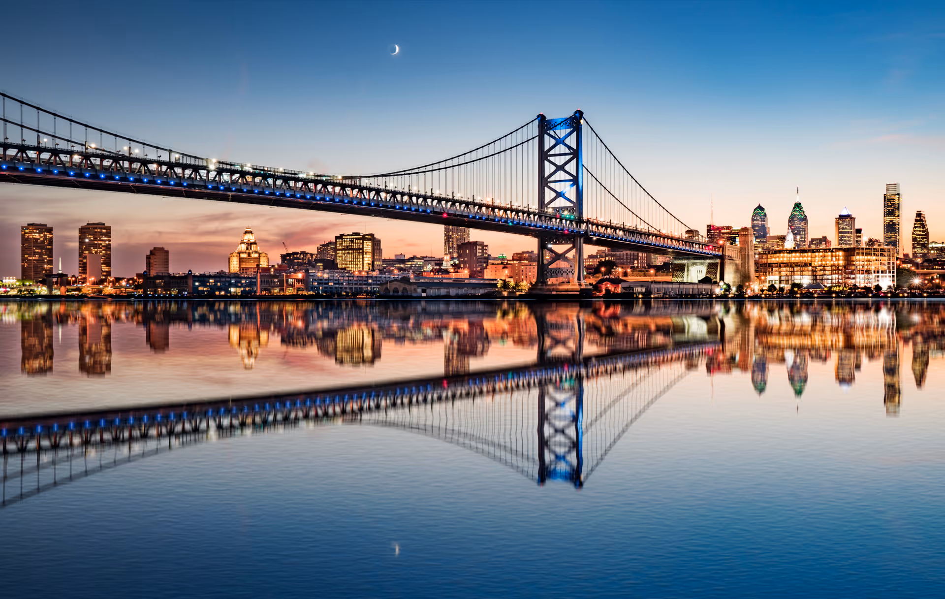 Nighttime city skyline with a large suspension bridge and its reflection in water.
