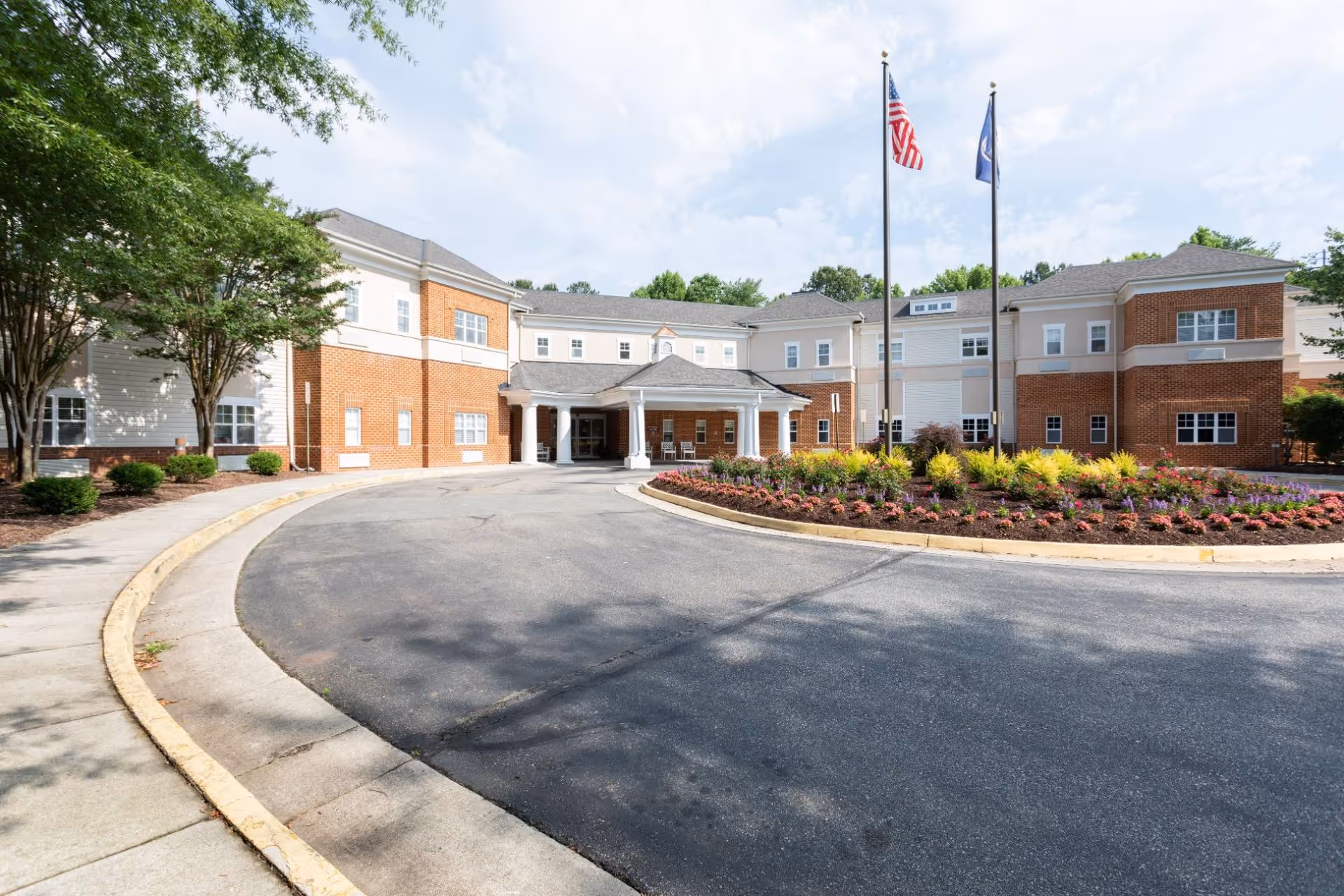 Front exterior view of a two-story senior living facility building with a circular driveway and a landscaped flower bed in the center. Two flagpoles with the American flag and another flag are visible near the entrance. Trees and shrubs surround the building under a partly cloudy sky.
