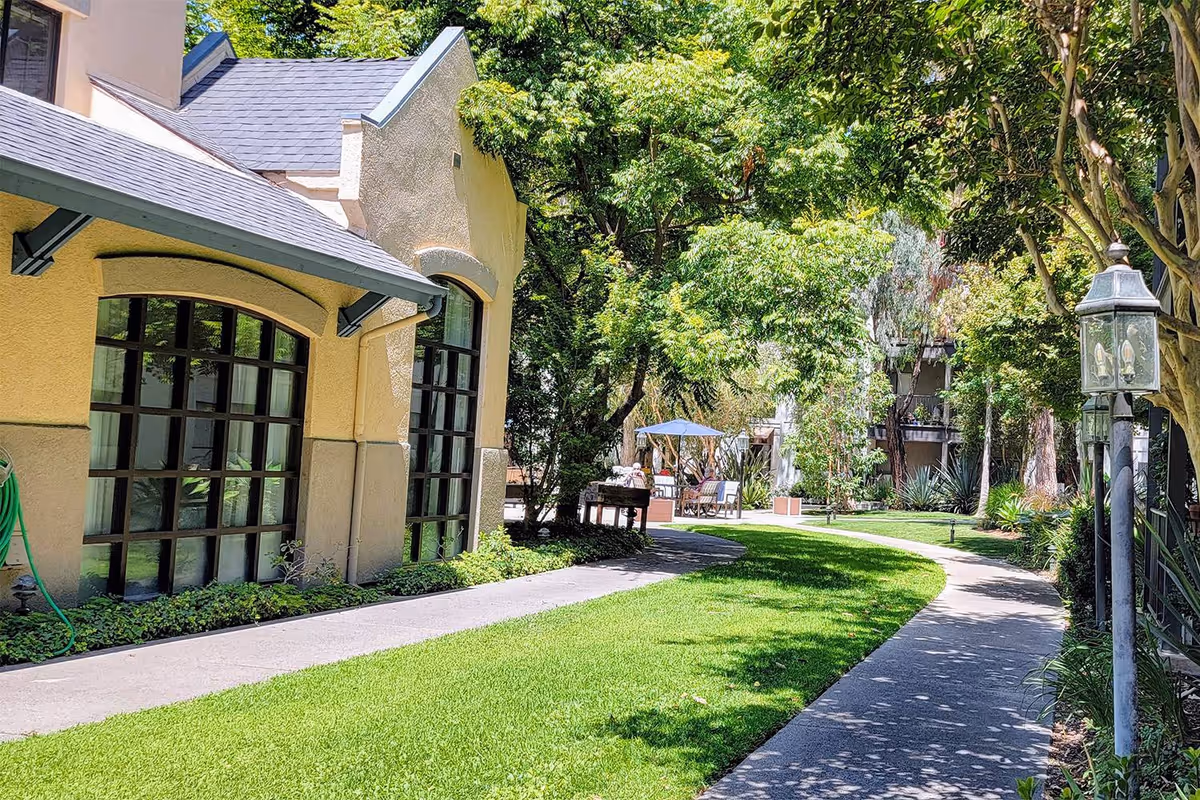 A sunny outdoor garden area at Westminster Terrace featuring a curved concrete pathway bordered by green grass and trees. The left side shows a beige building with large windows, and the right side has vintage-style lamp posts along the path. In the background, there is outdoor seating with an umbrella and lush greenery.