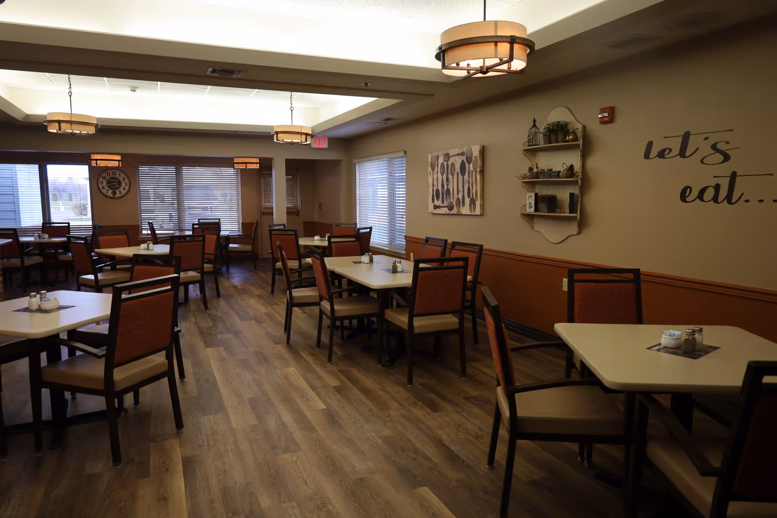 A dining room with multiple tables and chairs arranged neatly. The room has wooden flooring, beige walls with a brown lower trim, and large windows with blinds allowing natural light. Wall decorations include a clock, a painting of utensils, a small shelf with decorative items, and the phrase 'let's eat...' written on the wall. Ceiling lights provide warm lighting.