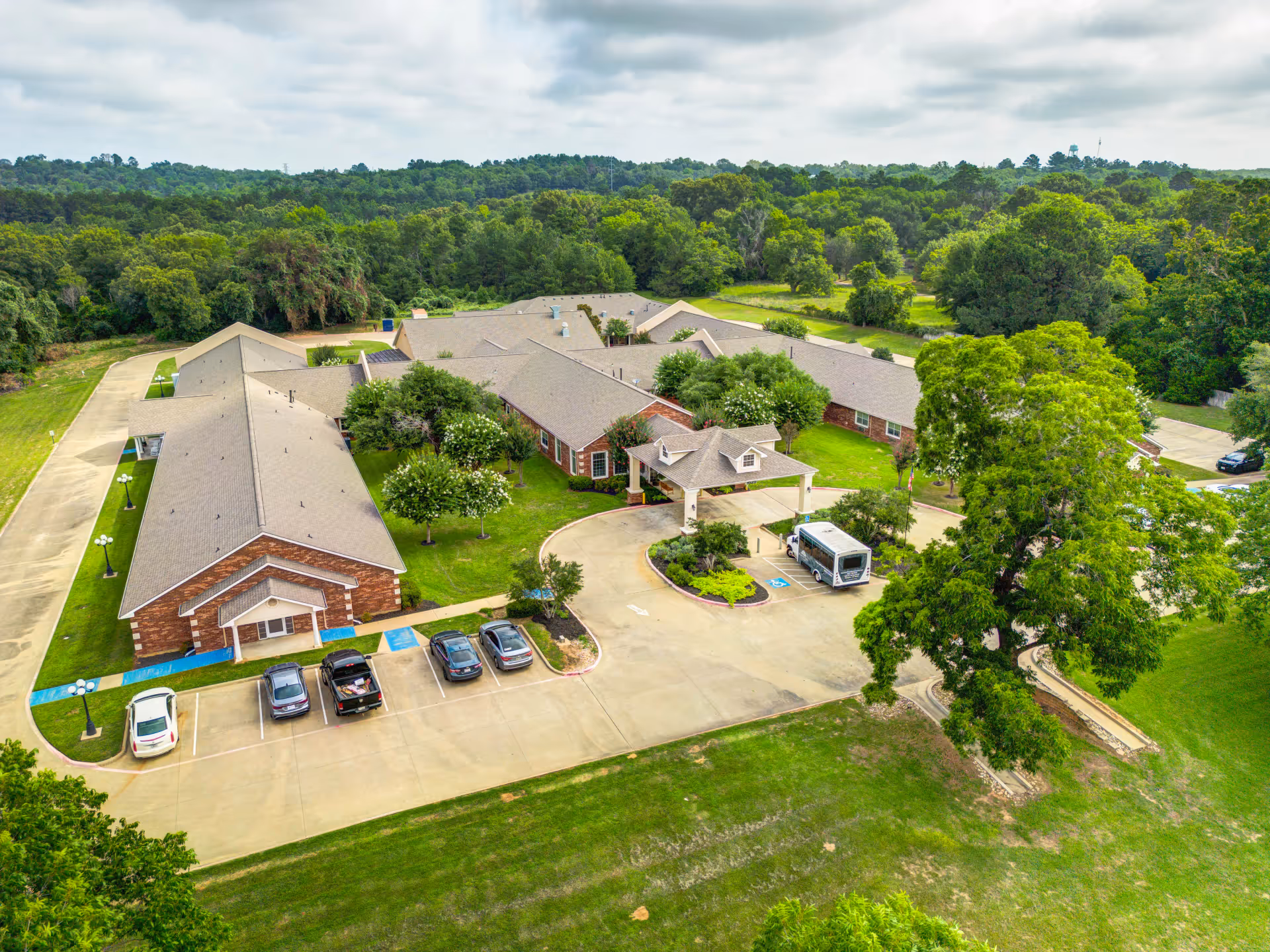 Aerial view of Dogwood Trails Assisted Living and Memory Care facility showing a large single-story brick building with multiple wings, surrounded by green lawns and trees. There is a circular driveway with a covered entrance, several parked cars, and a small shuttle bus. The background features a wooded area under a cloudy sky.