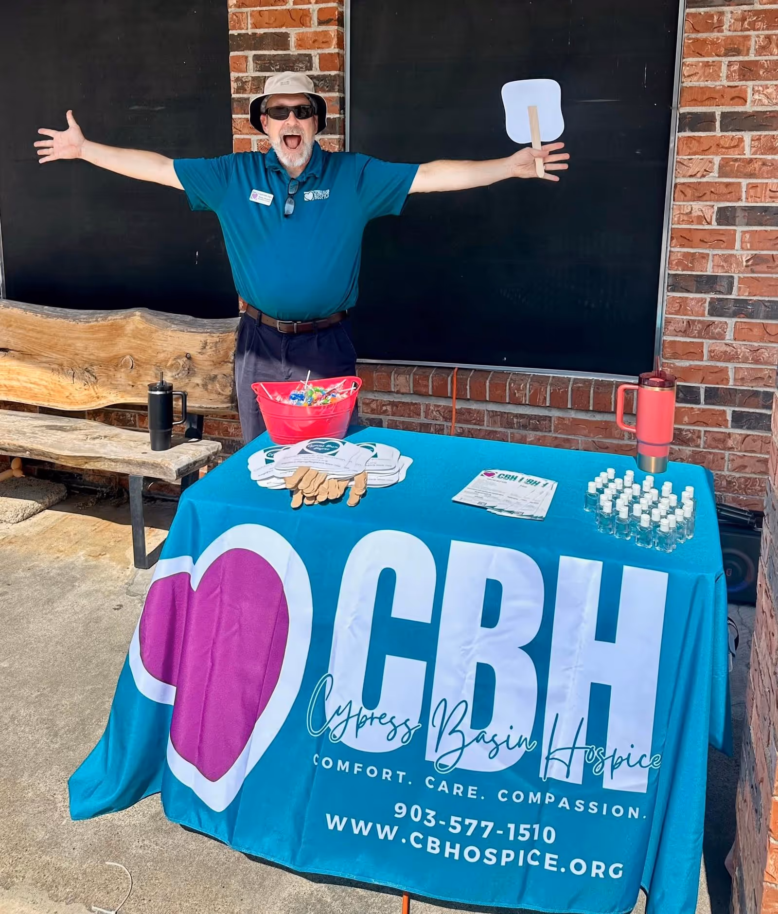 Smiling man in teal polo, hat and sunglasses, stands beside a blue bannered table outdoors.