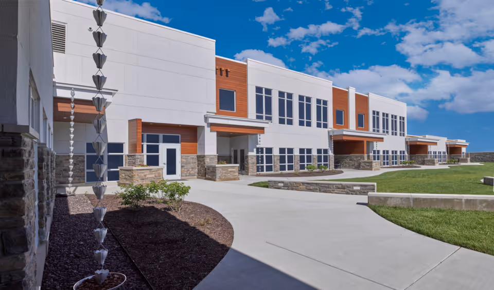 A modern two-story senior living facility exterior with white and wood accents along a curved walkway.