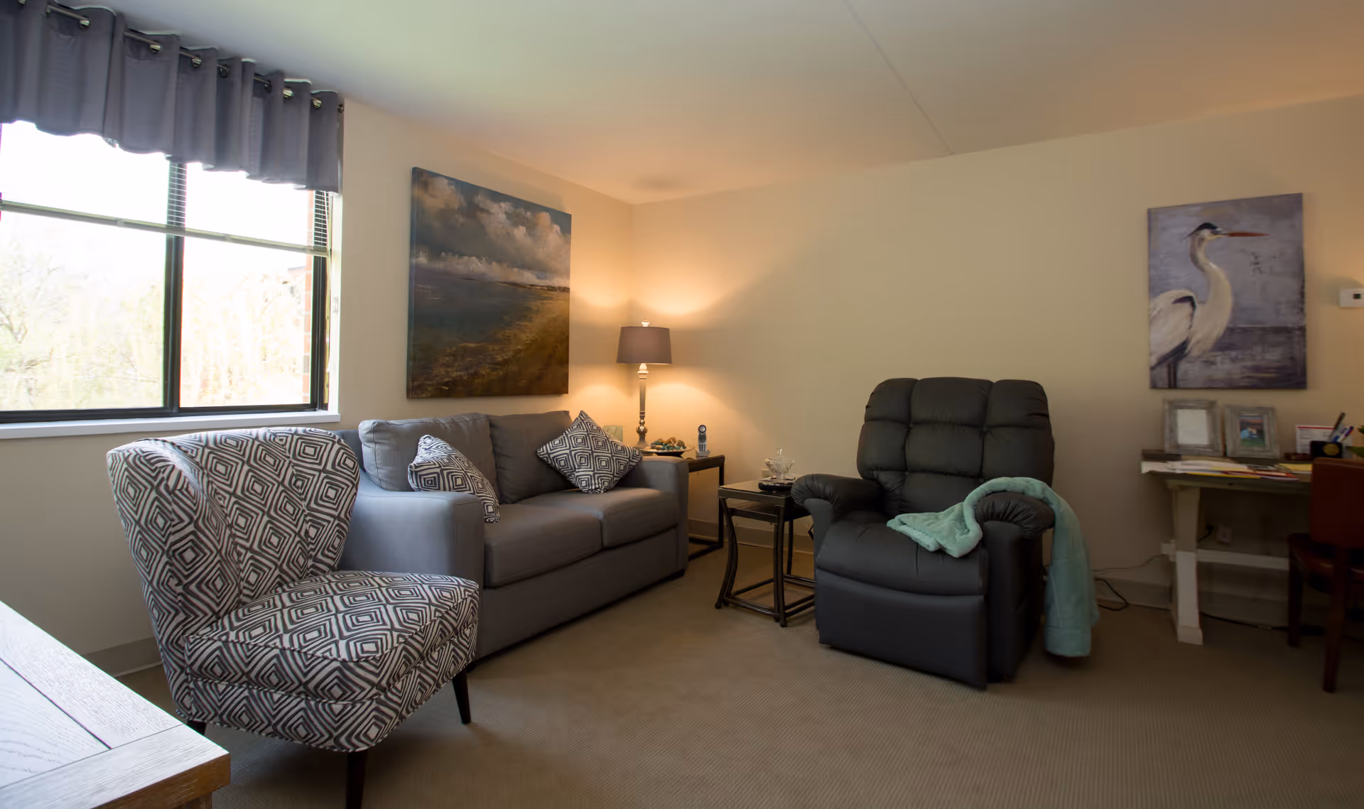 Living room with a patterned armchair, gray sofa, recliner, side tables, lamp and wall art.