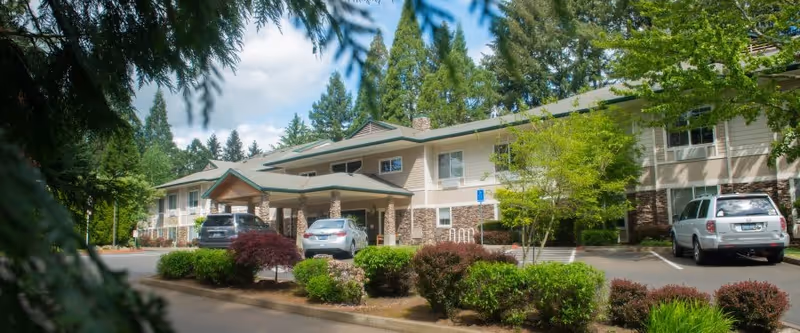 Exterior front of a two-story assisted living facility with a covered entrance, parked cars, and landscaped grounds.