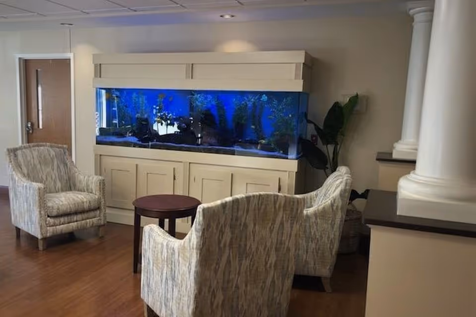 Upholstered chairs arranged around a small round table facing a large built-in aquarium in a communal seating area.