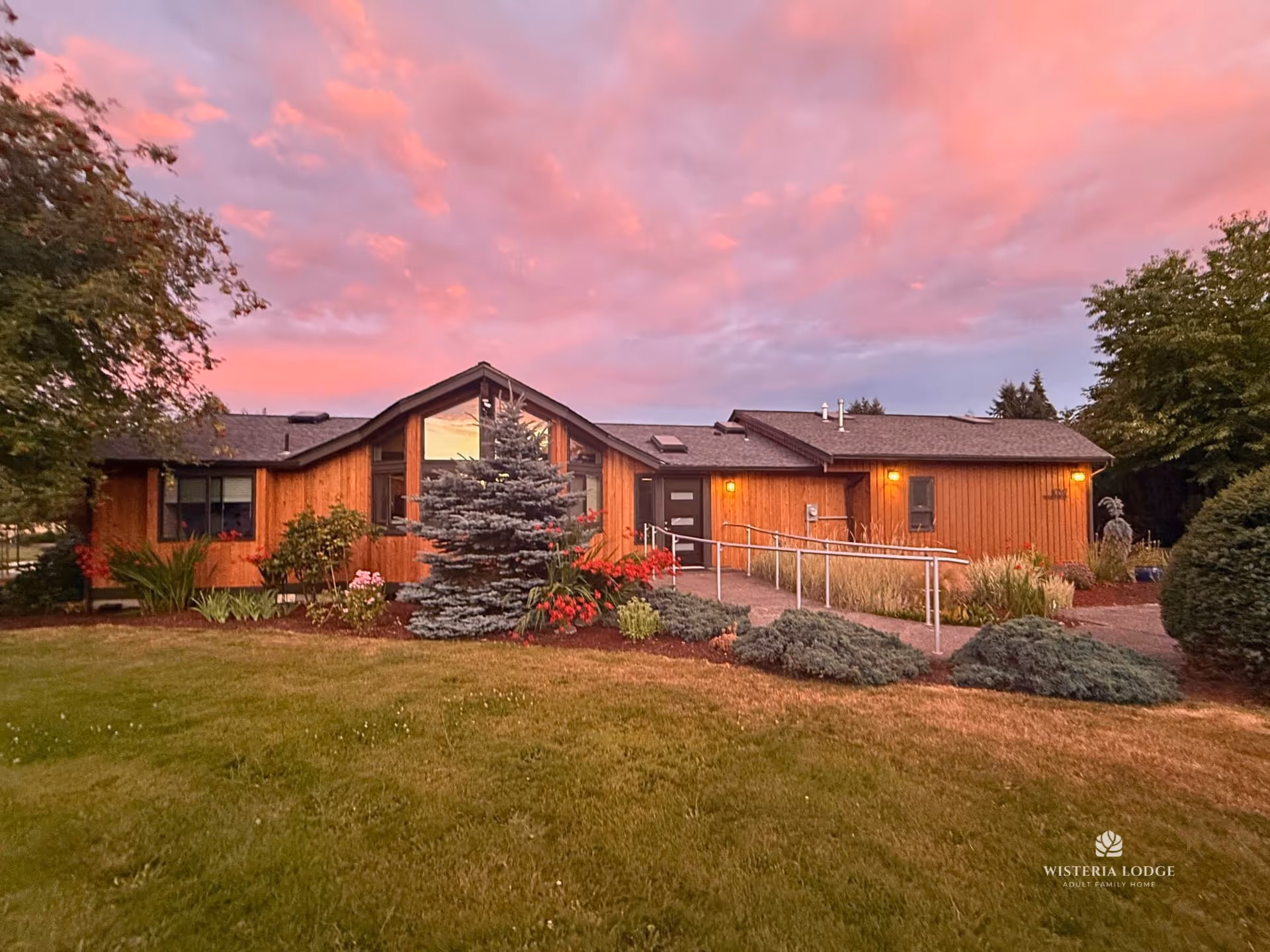Wood-clad senior living facility with ramp, landscaped front yard, and pink sunset sky.