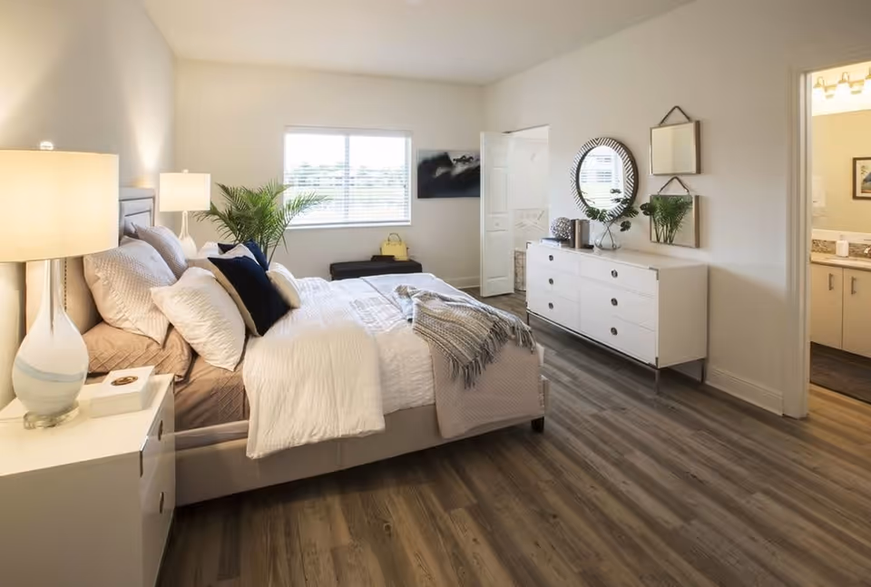Bright, modern bedroom with a made bed, nightstand and dresser, a window and doorway to an en-suite bathroom.