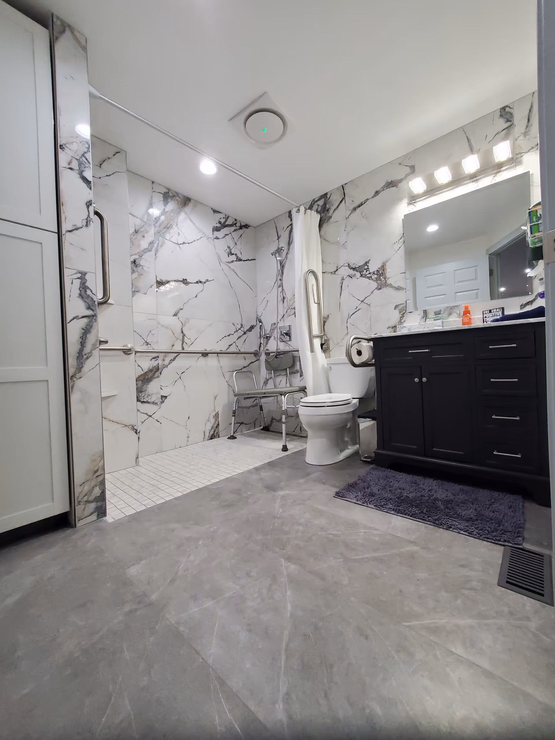 Accessible bathroom featuring marble walls, a shower, toilet, and dark vanity.