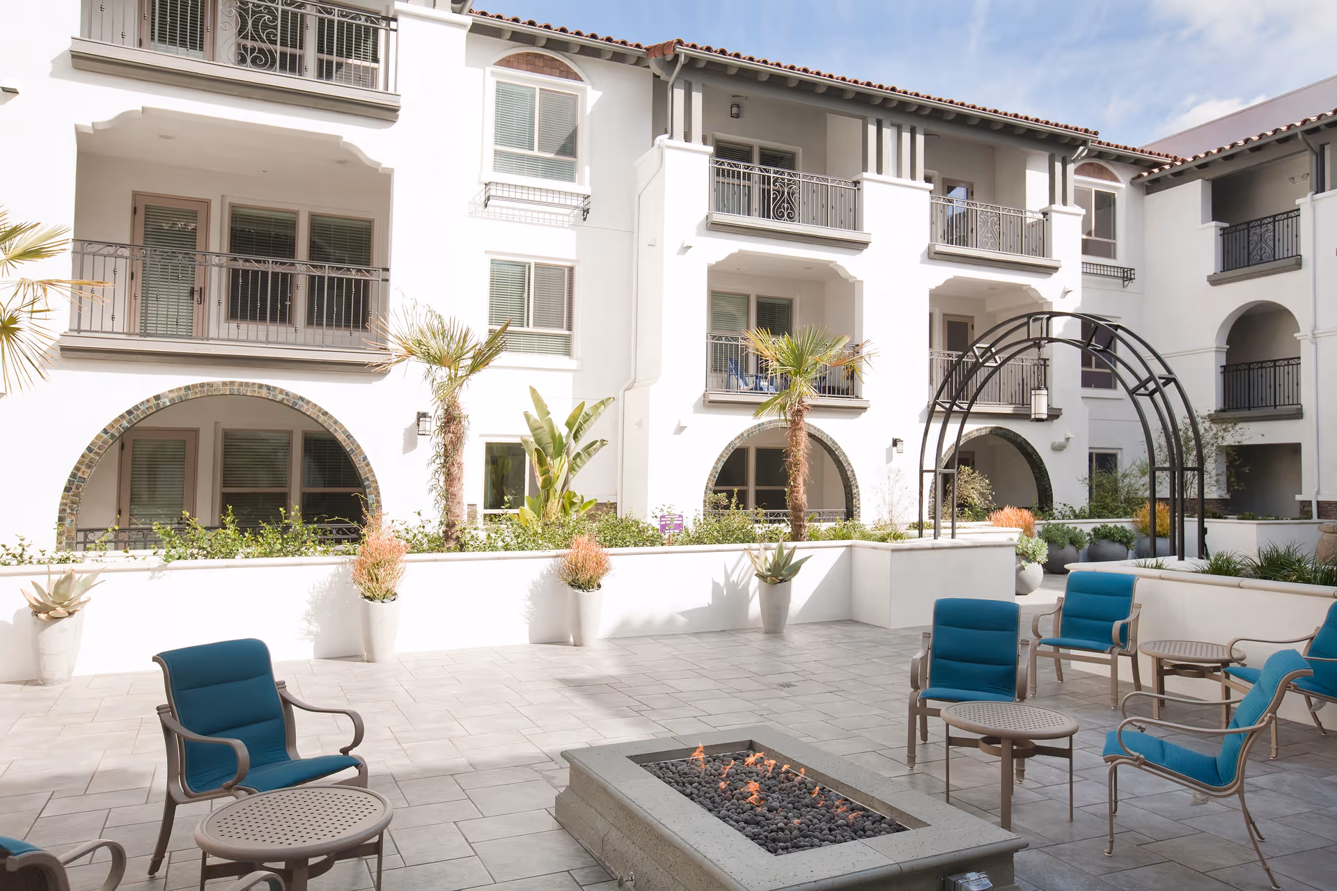 Outdoor courtyard area of a senior living facility with a tiled floor, blue cushioned chairs arranged around a rectangular fire pit, potted plants along the walls, palm trees, and a white multi-story building with balconies and arched doorways in the background under a partly cloudy sky.