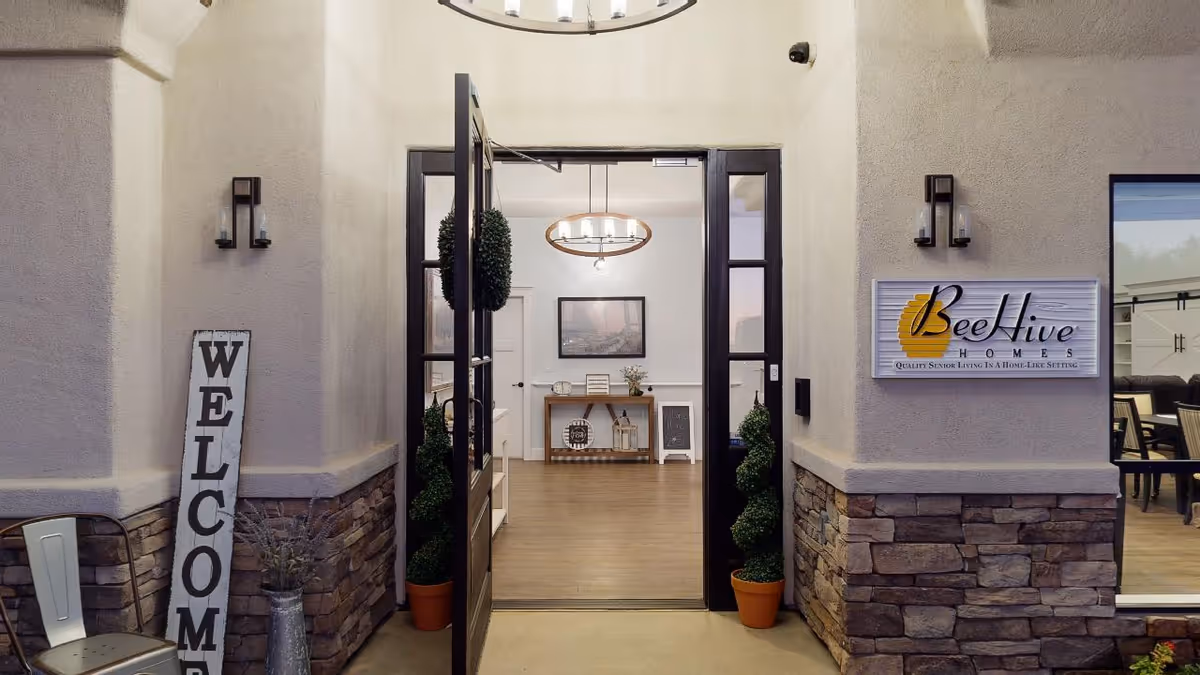 Entrance to BeeHive Homes Assisted Living facility with open double doors, two potted topiary plants on either side, a vertical welcome sign on the left, and a BeeHive Homes sign on the right. Inside, a wooden console table with decorative items and a framed picture on the wall are visible.