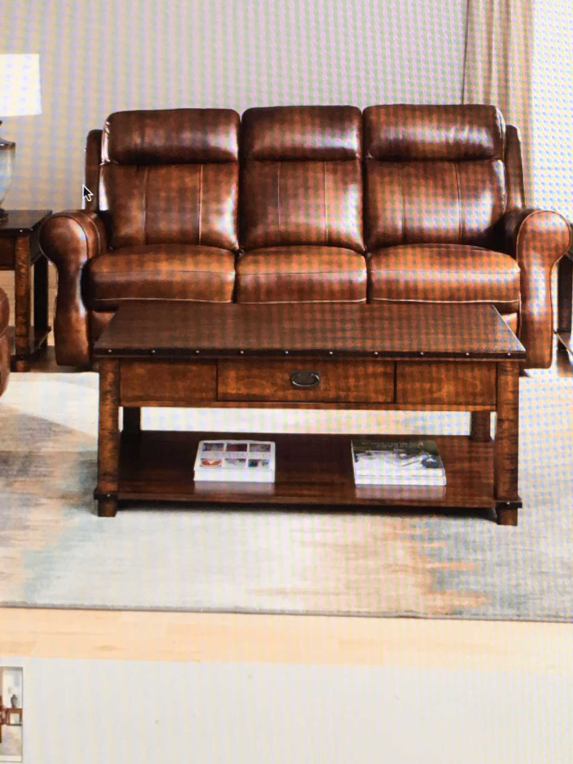 A brown leather three-seater sofa with a wooden coffee table in front of it. The coffee table has a drawer and a lower shelf with two books placed on it. The setting appears to be a living room with a light-colored rug underneath the furniture.