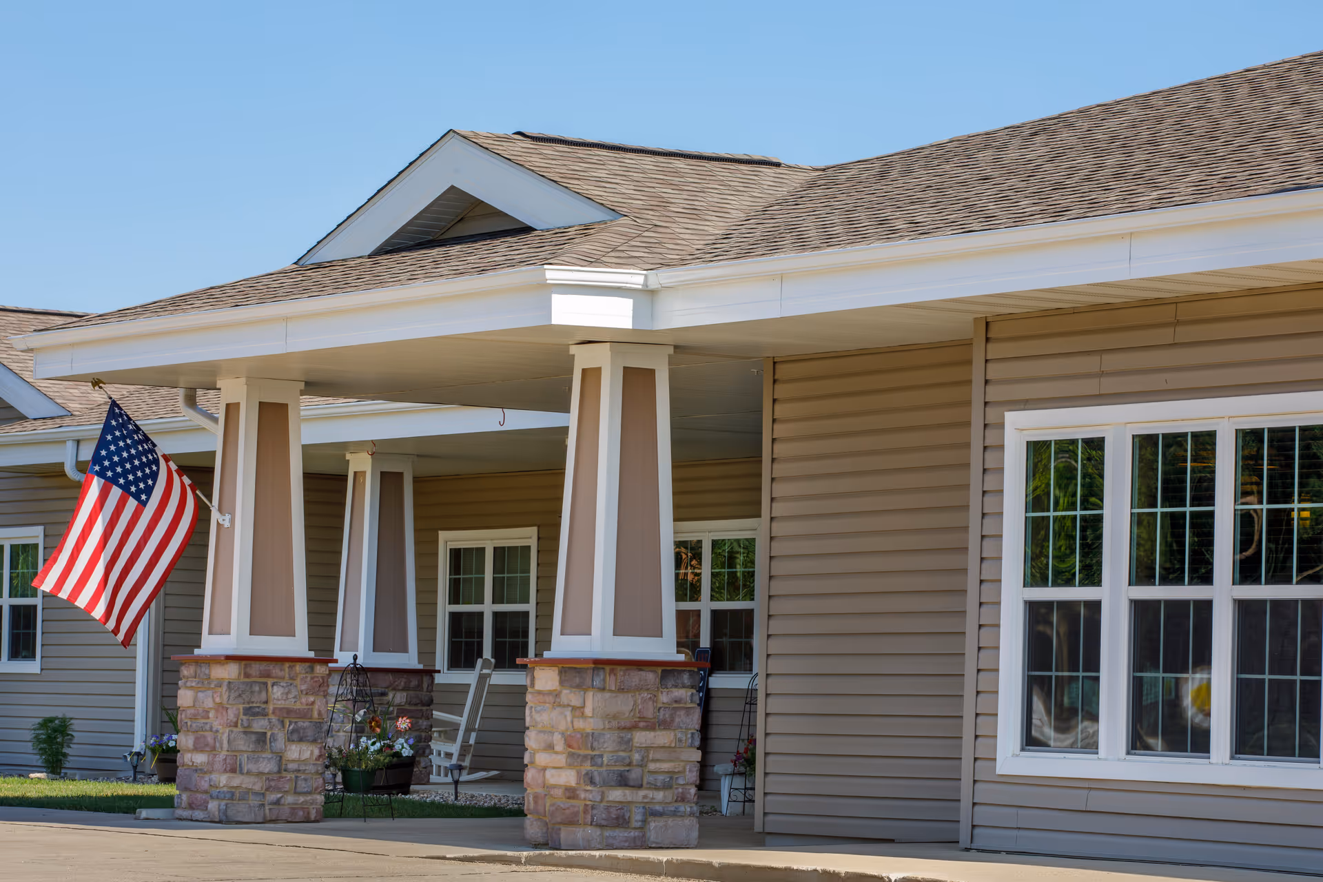 Exterior view of a building entrance with a covered porch supported by stone and wooden pillars, an American flag mounted on the left side, and windows with white frames on the beige siding walls.