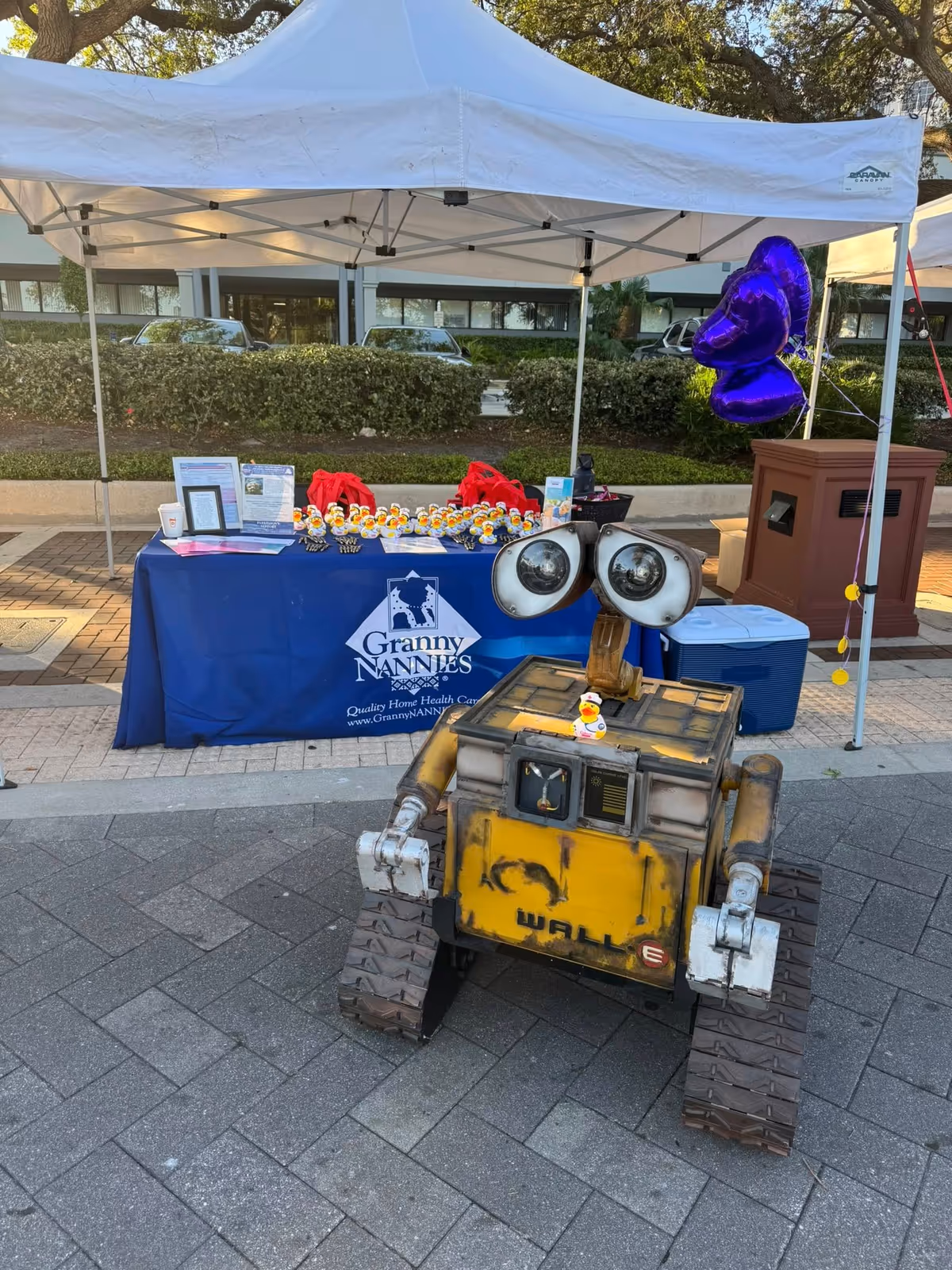 Outdoor event booth with Granny NANNIES table, blue cover, and WALL-E style robot.