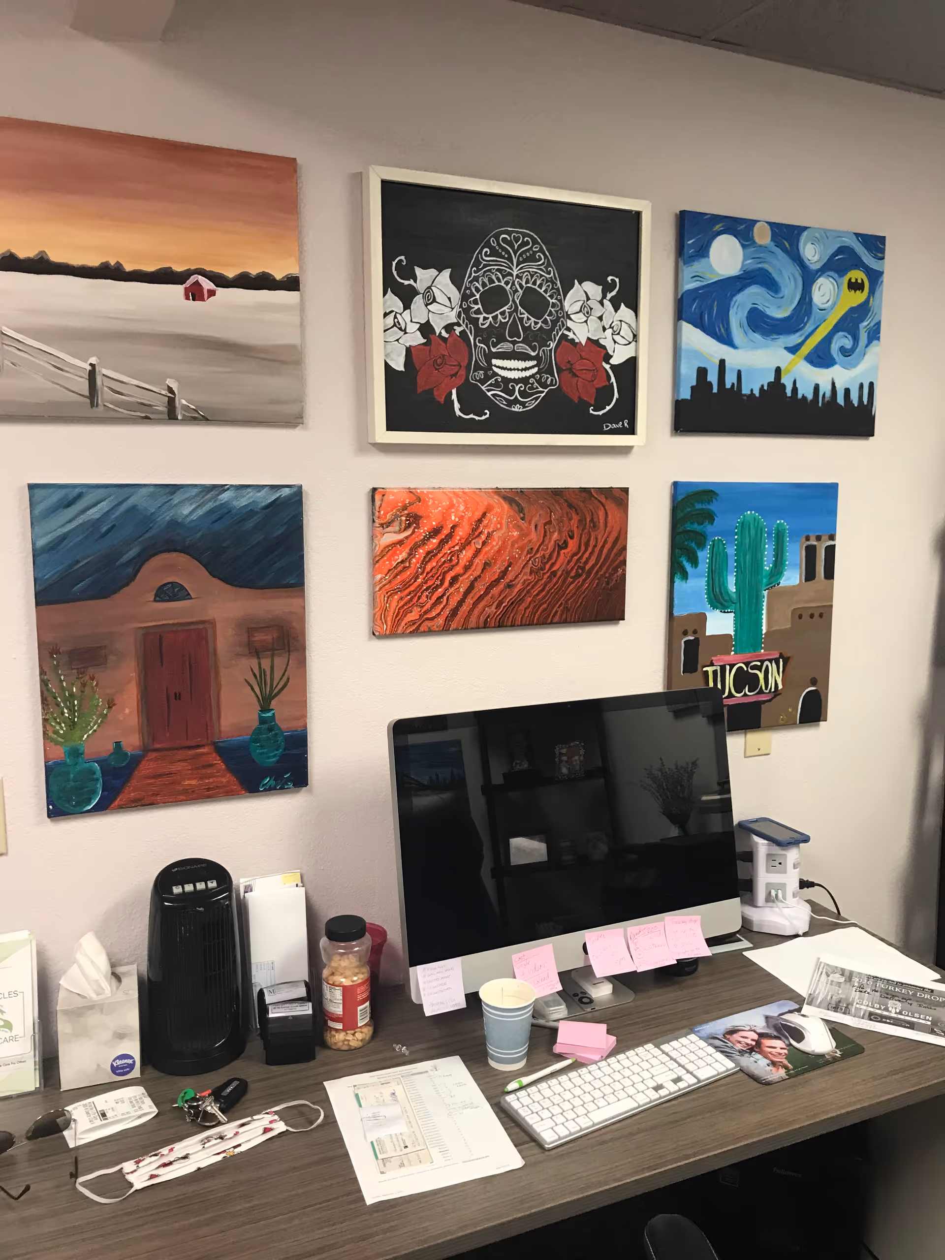 An office desk against a wall of colorful art prints with computer and supplies.