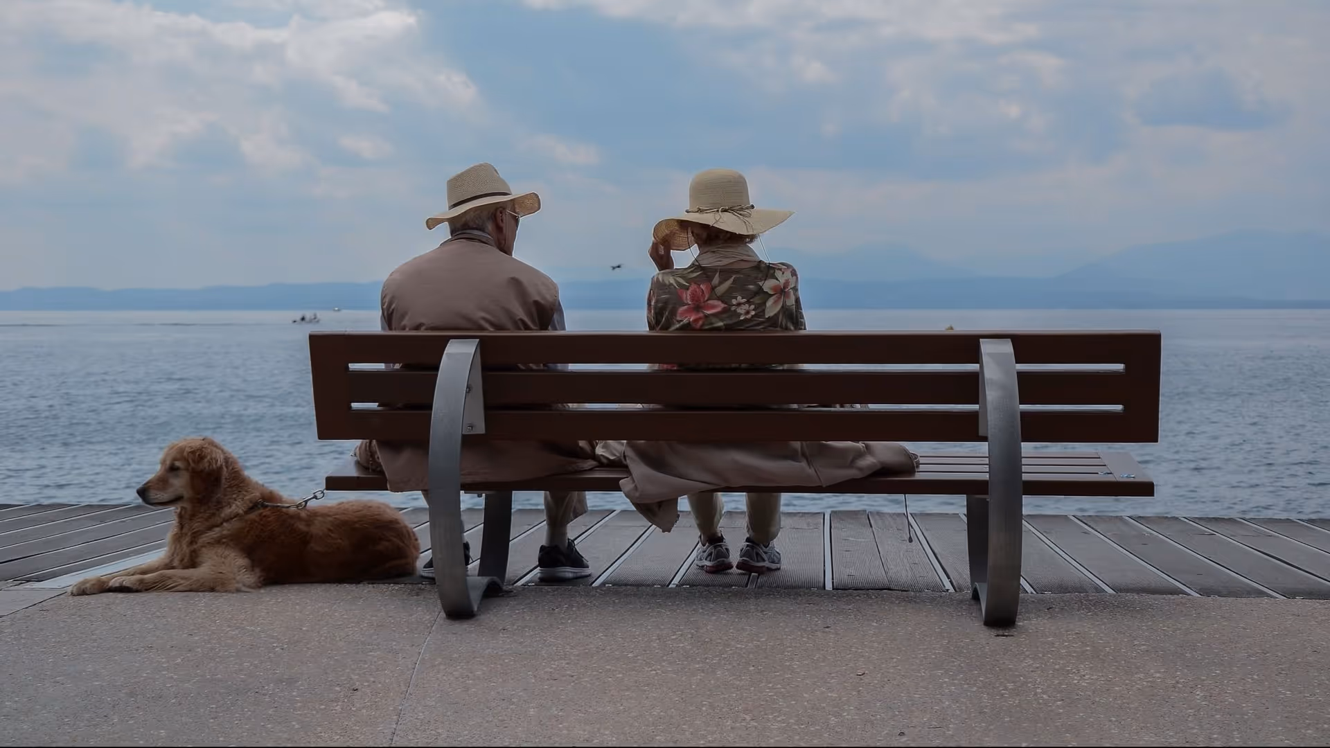 Two seniors sit on a bench by a lake with a leashed dog nearby.