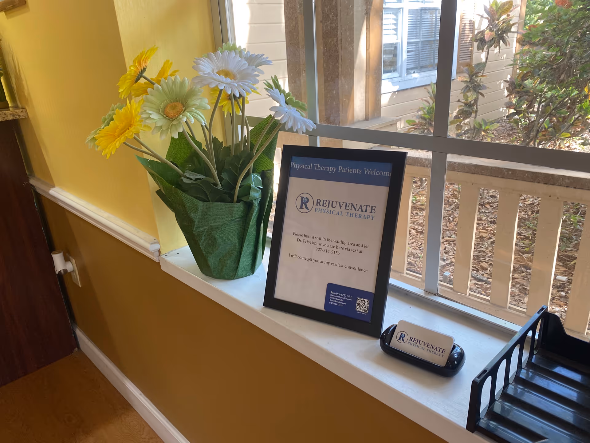 Yellow-walled interior with a windowsill display of flowers and a framed therapy notice.