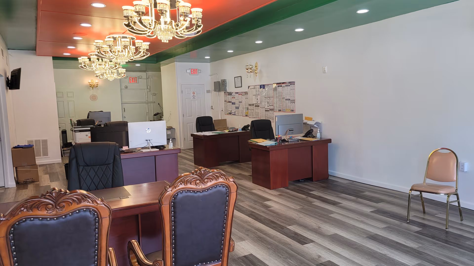 Interior office reception area with desks, computers, chandeliers, and an empty chair along the wall.
