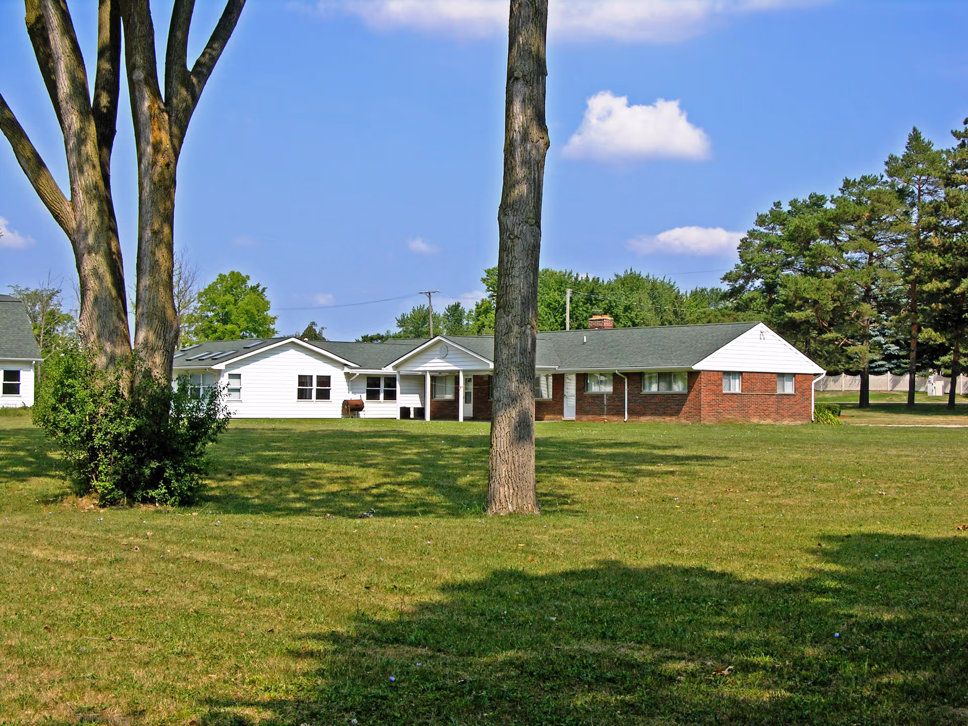Single-story building with white siding and red brick exterior surrounded by a large grassy lawn with trees under a blue sky with a few clouds.
