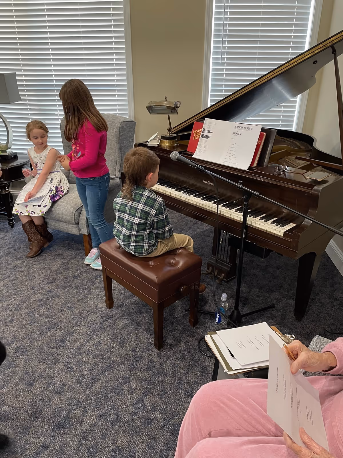 Children and an older adult gathered around a grand piano in a carpeted common room, with one child seated at the piano bench.