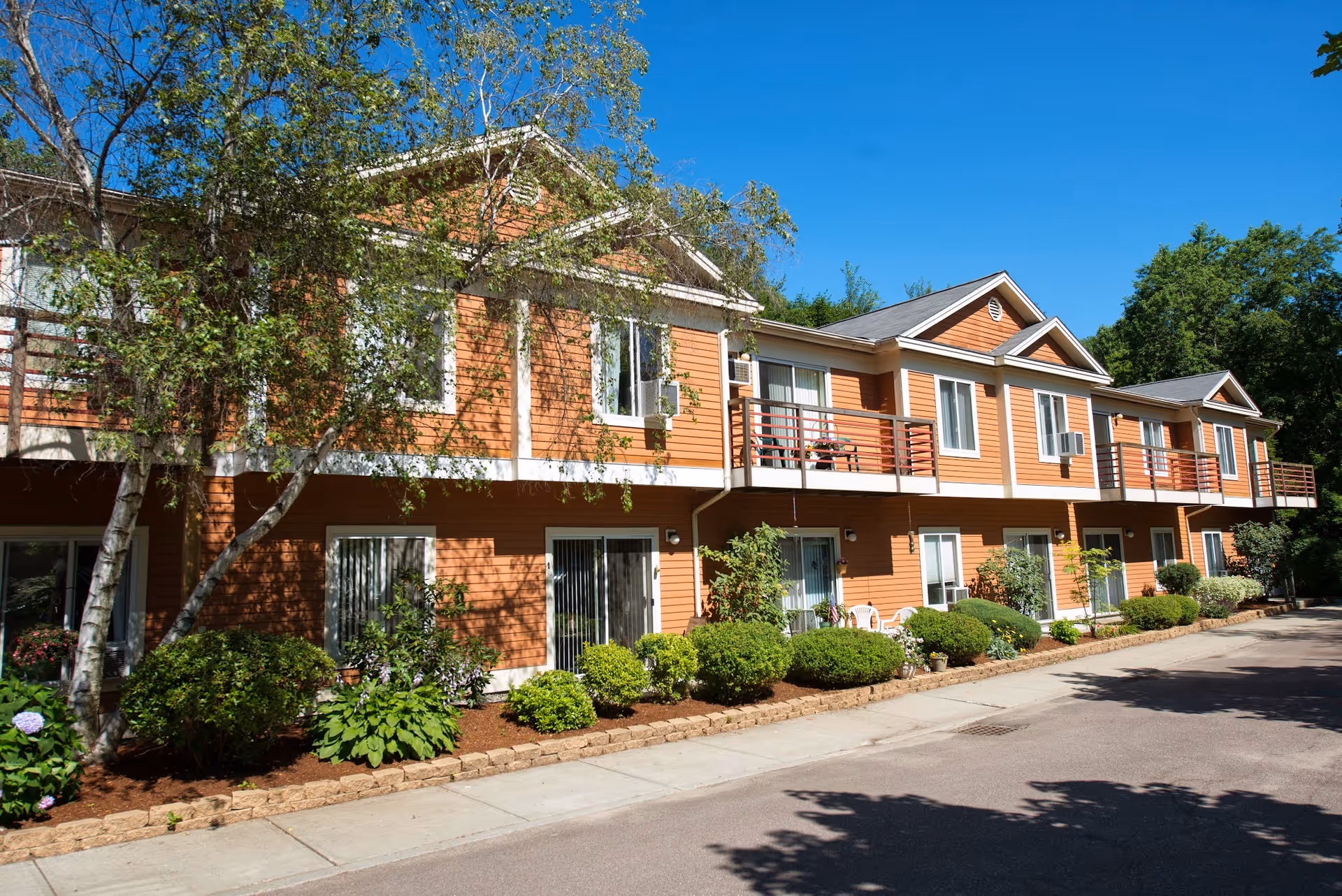 Two-story orange senior living facility with balconies, shrubs, and a paved entrance walkway.