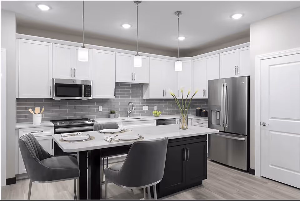 Modern kitchen with white upper cabinets, gray subway tile backsplash, stainless steel appliances including a microwave, stove, and refrigerator. A large island with a white countertop and dark base has two gray upholstered chairs and is set with plates, napkins, and cutlery. Three pendant lights hang above the island, and a vase with white flowers is placed on the island.