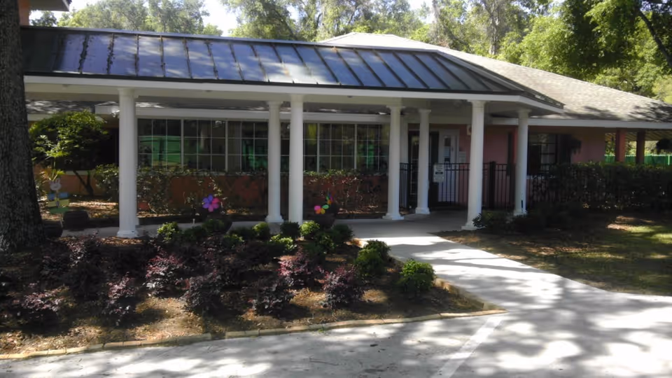 Covered entrance with white columns and a walkway leading to the front doors of a one-story assisted living building surrounded by landscaping.