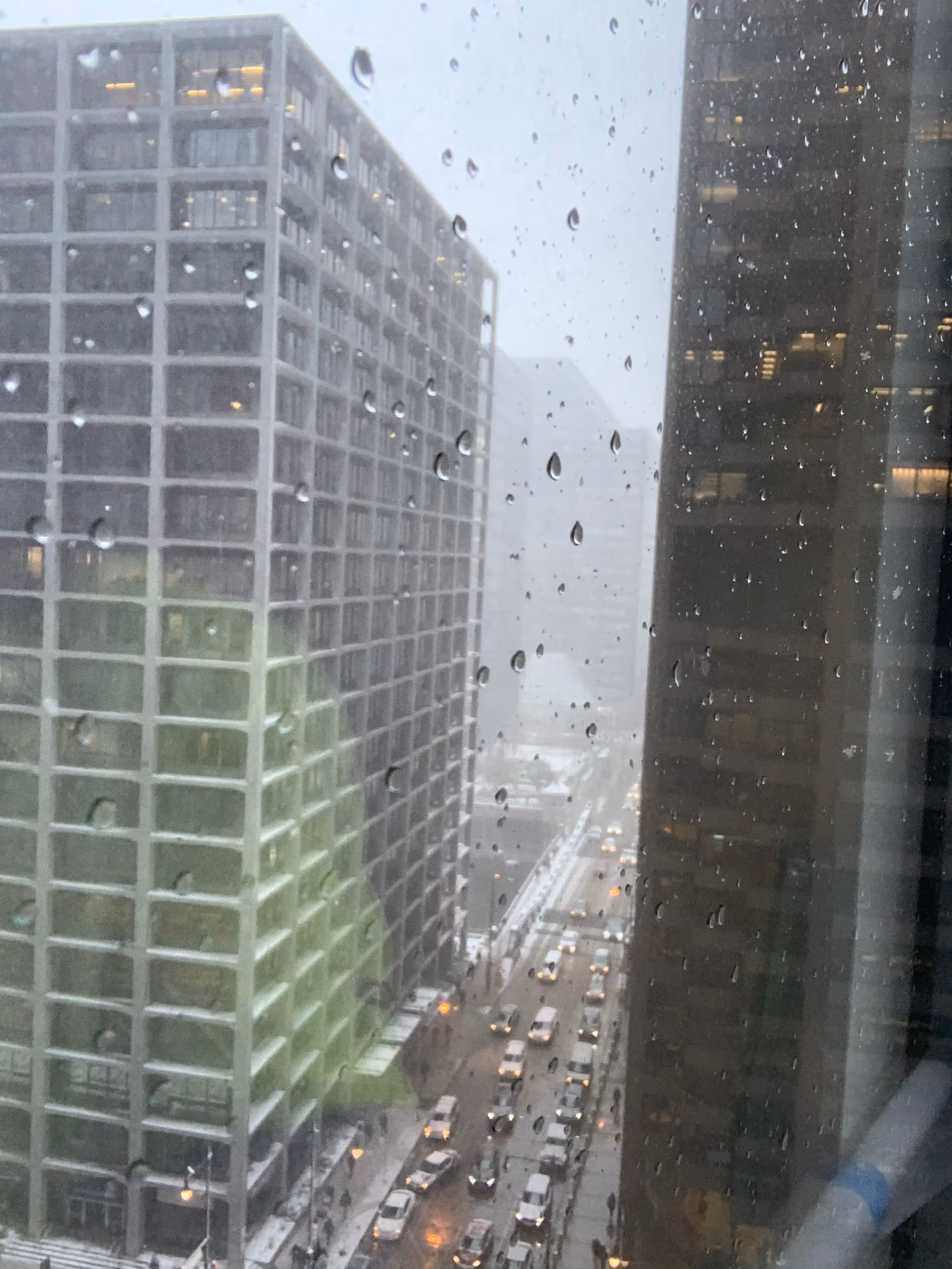 Rain-spattered window view of tall city buildings and a busy street below.