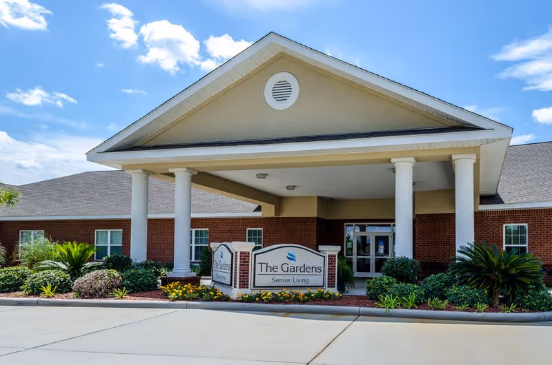 Exterior view of The Gardens Senior Living entrance with pillars and landscaped walkway