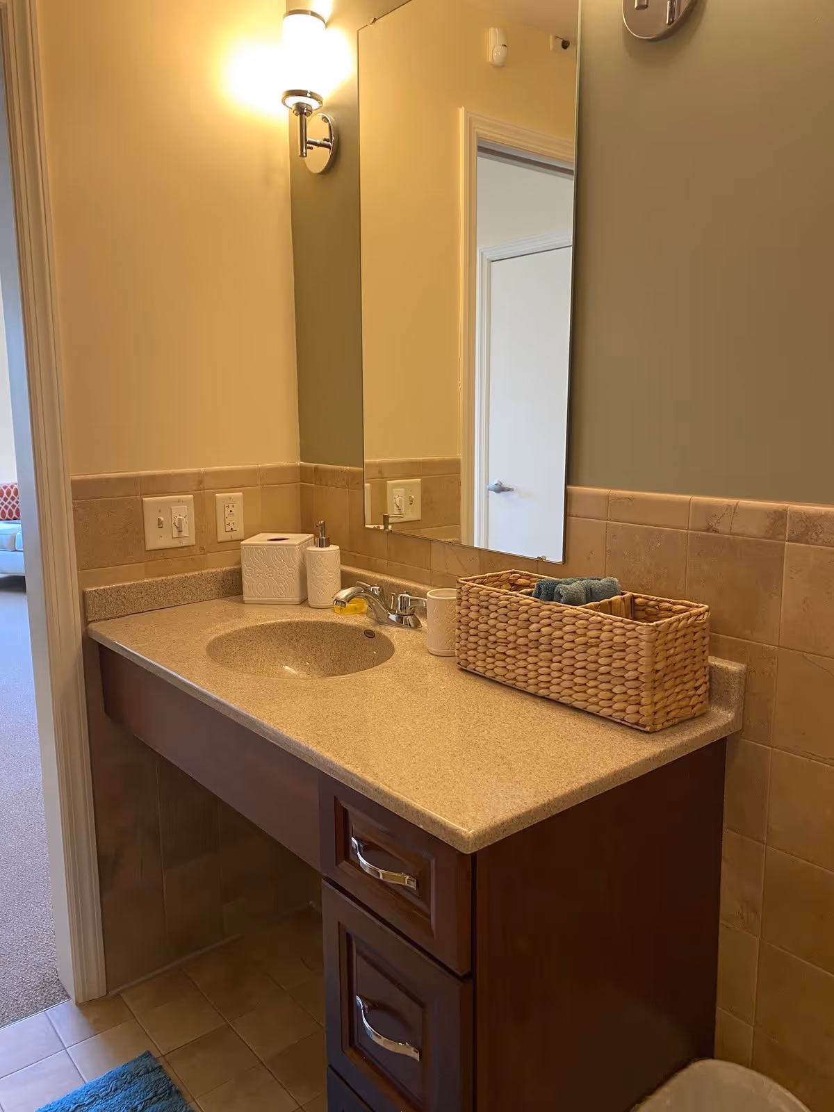 A bathroom vanity with a beige countertop and an inset sink. On the countertop, there is a woven basket containing towels, a soap dispenser, a tissue box, and a cup. Above the vanity is a rectangular mirror and a wall-mounted light fixture. The walls are painted in two tones, beige tiles halfway up and greenish paint above. The bathroom floor is tiled, and a blue bath mat is partially visible. A doorway leads to a carpeted room with a sofa visible in the background.