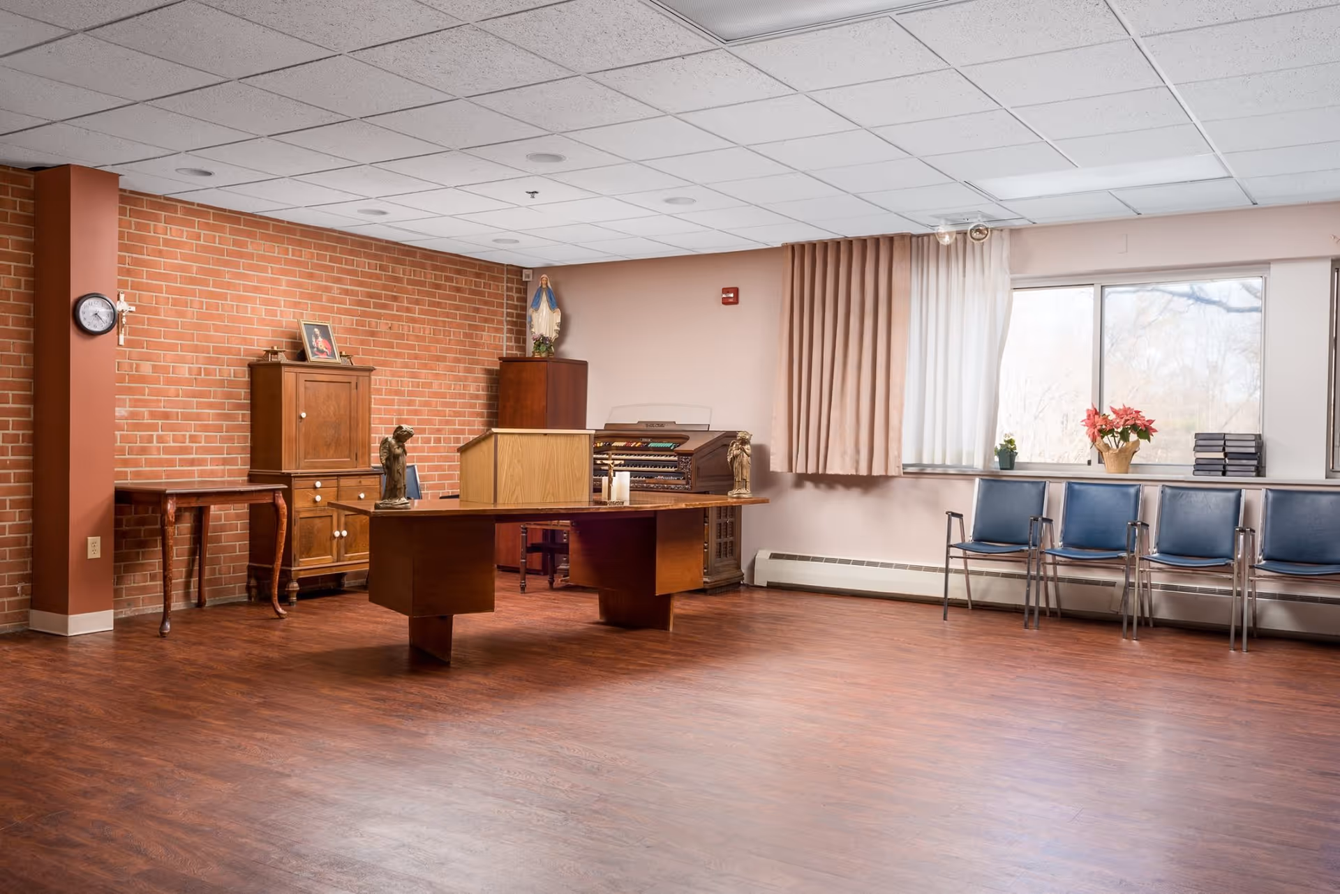 A spacious room with wooden flooring and a brick accent wall. The room contains a wooden table with religious statues and candles, a wooden cabinet, an organ, and a row of blue chairs along the window. The window has beige curtains and there are plants and flowers on the windowsill.