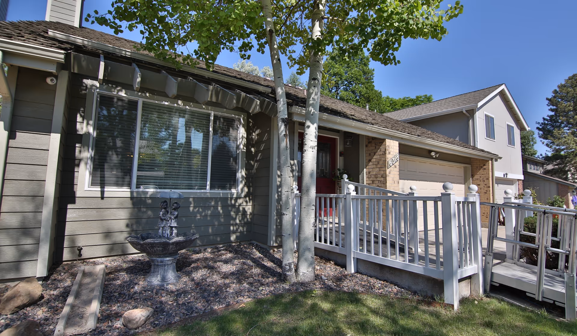 Front exterior of an assisted living residence showing a porch with white ramp railing, garage, window, small rock garden with a fountain, and trees.