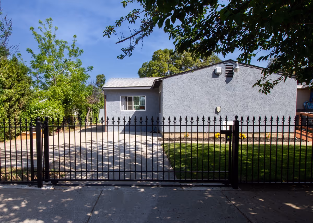 View of a single-story gray house with a white roof behind a black metal fence. There is a concrete driveway and a small green lawn with some yellow flowers. Trees and bushes surround the property under a clear blue sky.