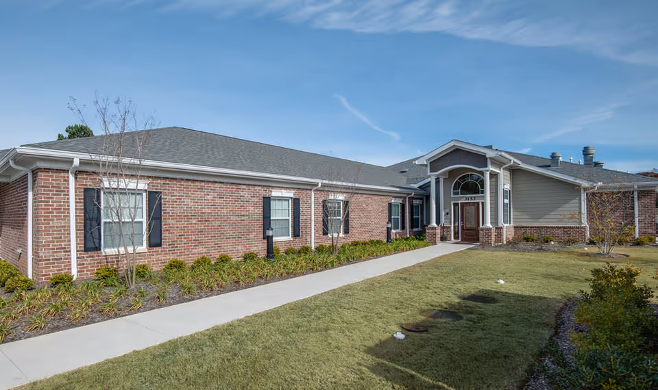 Exterior view of a single-story brick building with a gray shingled roof, white gutters, and black window shutters. A concrete walkway leads to a covered entrance with a wooden door and large windows above it. The building is surrounded by a well-maintained lawn and small shrubs under a clear blue sky.