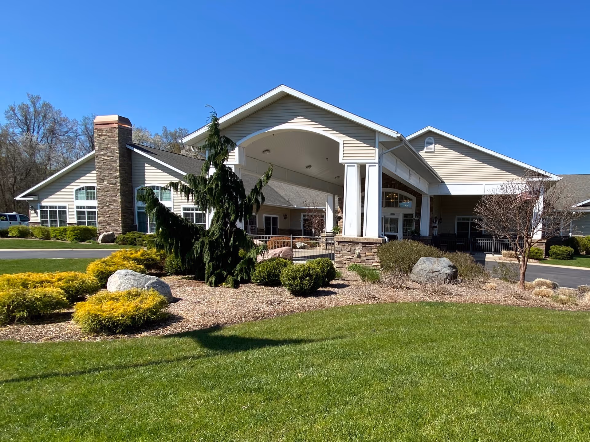 Front entrance of a single-story assisted living building with a covered porte-cochere, landscaped shrubs and lawn under a clear blue sky.