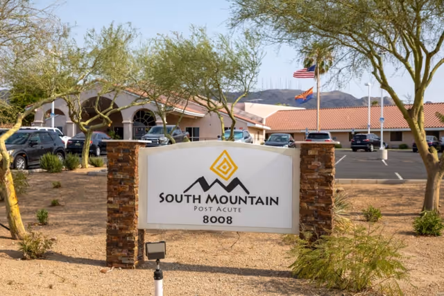 Outdoor view of South Mountain Post Acute facility sign with the building and parking lot in the background, surrounded by desert landscaping and trees under a clear sky.