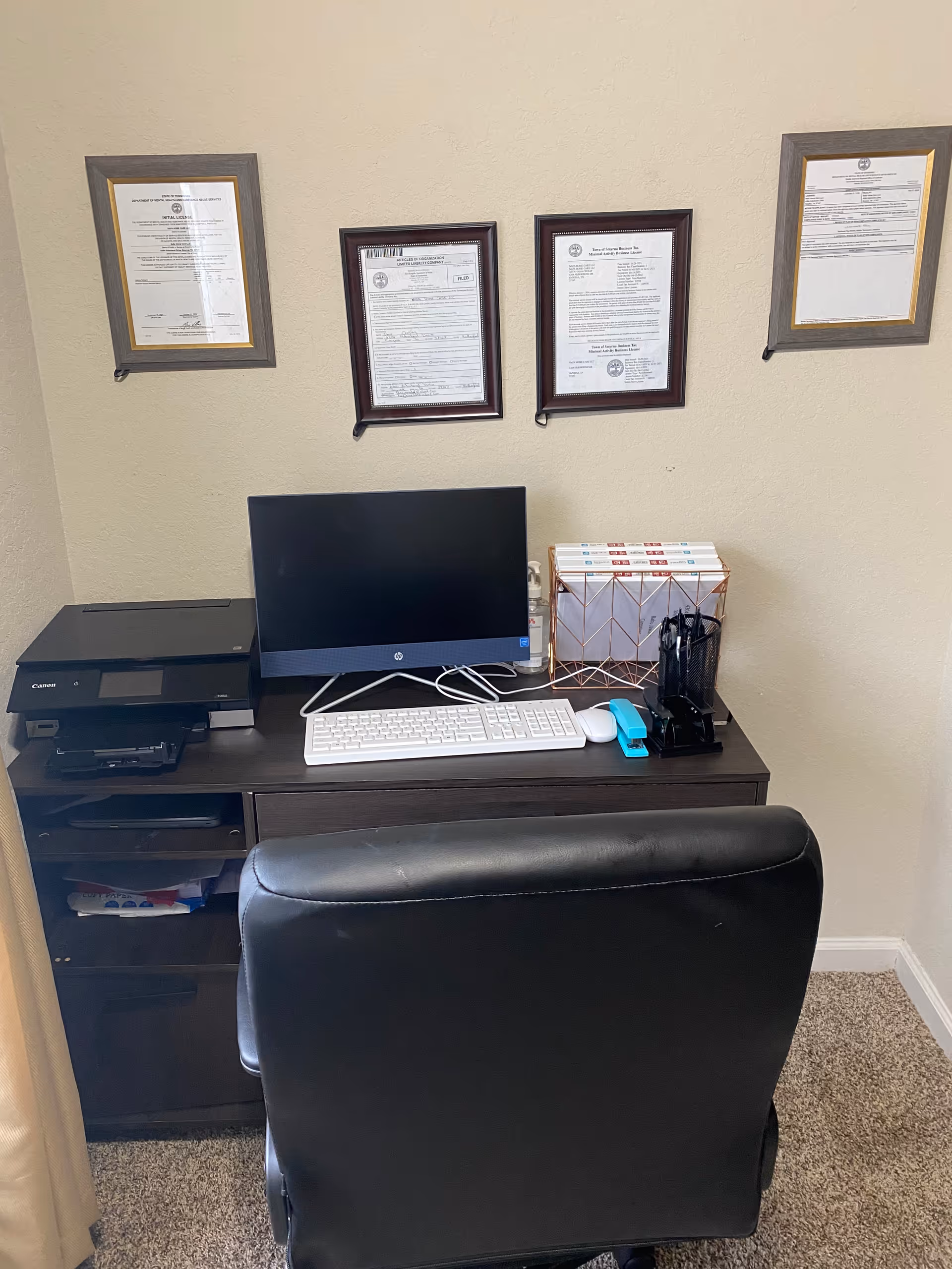 Office workspace with desktop computer, printer, keyboard, and framed documents on beige wall.