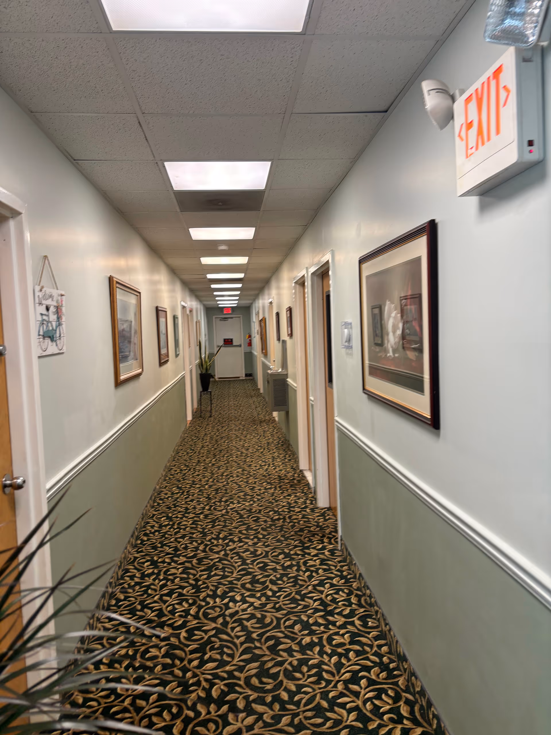 Long, carpeted interior hallway with framed art, doors on both sides, and an exit sign.