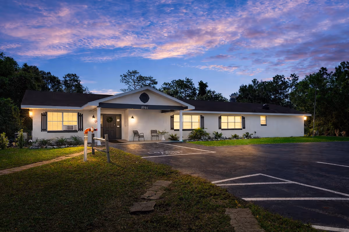 Single-story white senior living facility with front entrance and parking lot at dusk