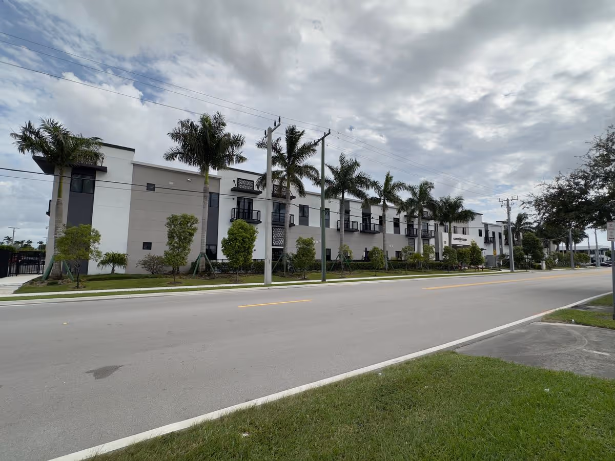 Street view of a modern three-story building with balconies and palm trees lining the sidewalk under a cloudy sky.