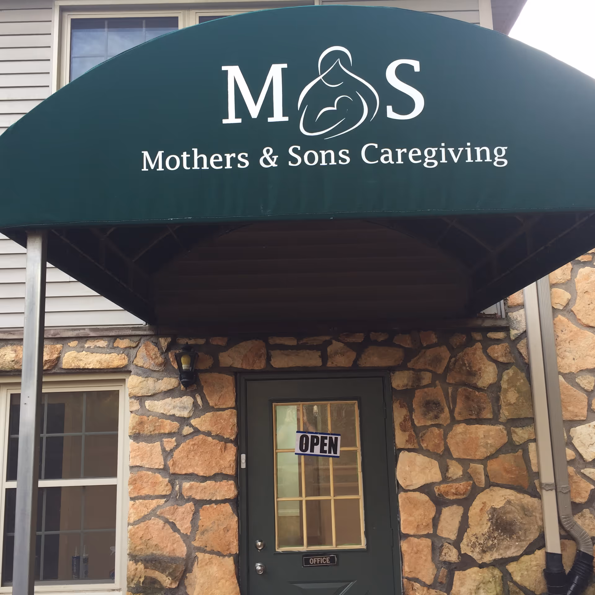 Stone-faced building entrance with green awning displaying Mothers and Sons Caregiving and Open sign.