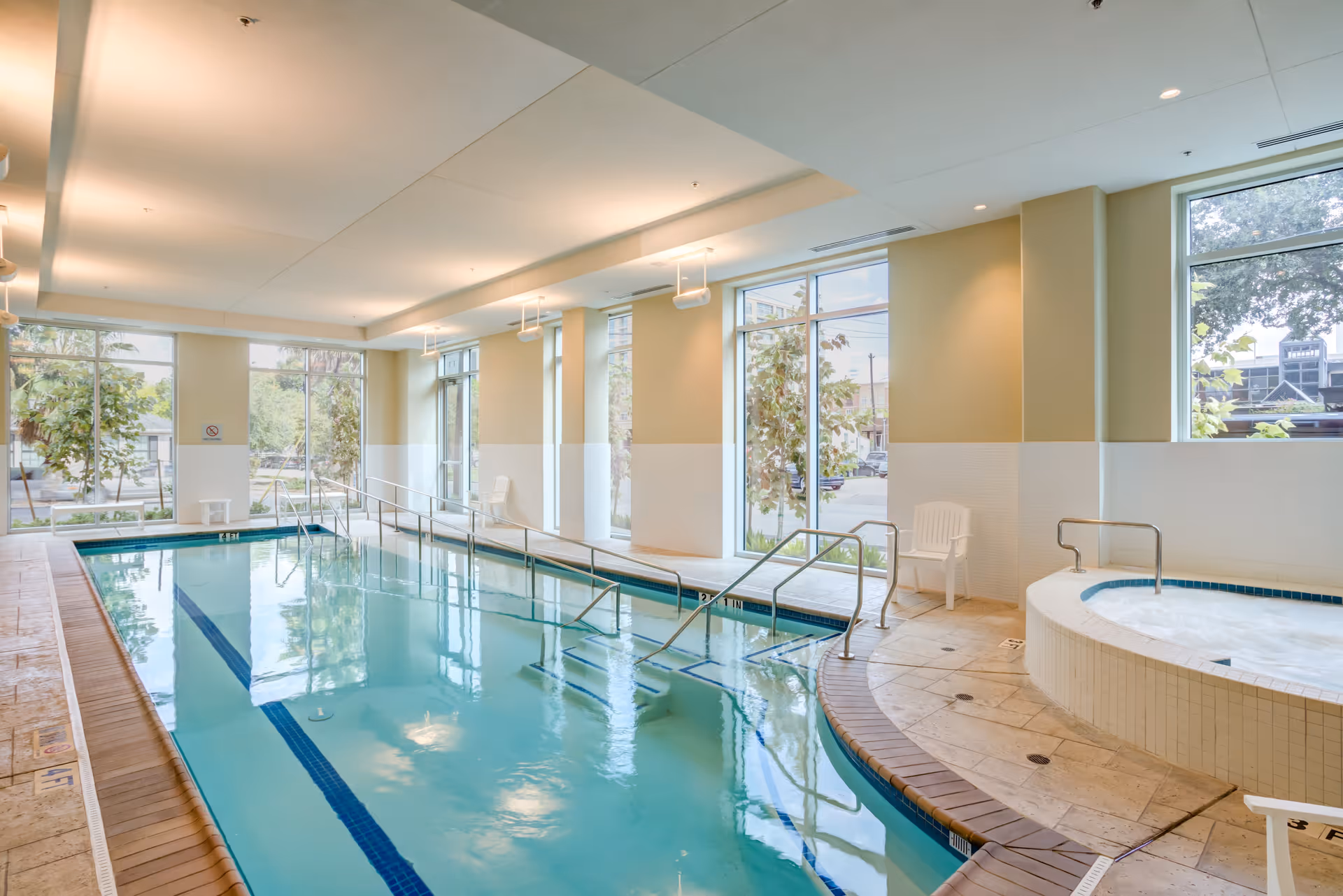 Indoor swimming pool area with clear blue water, handrails for easy access, large windows letting in natural light, and a round hot tub on the right side with white chairs nearby.