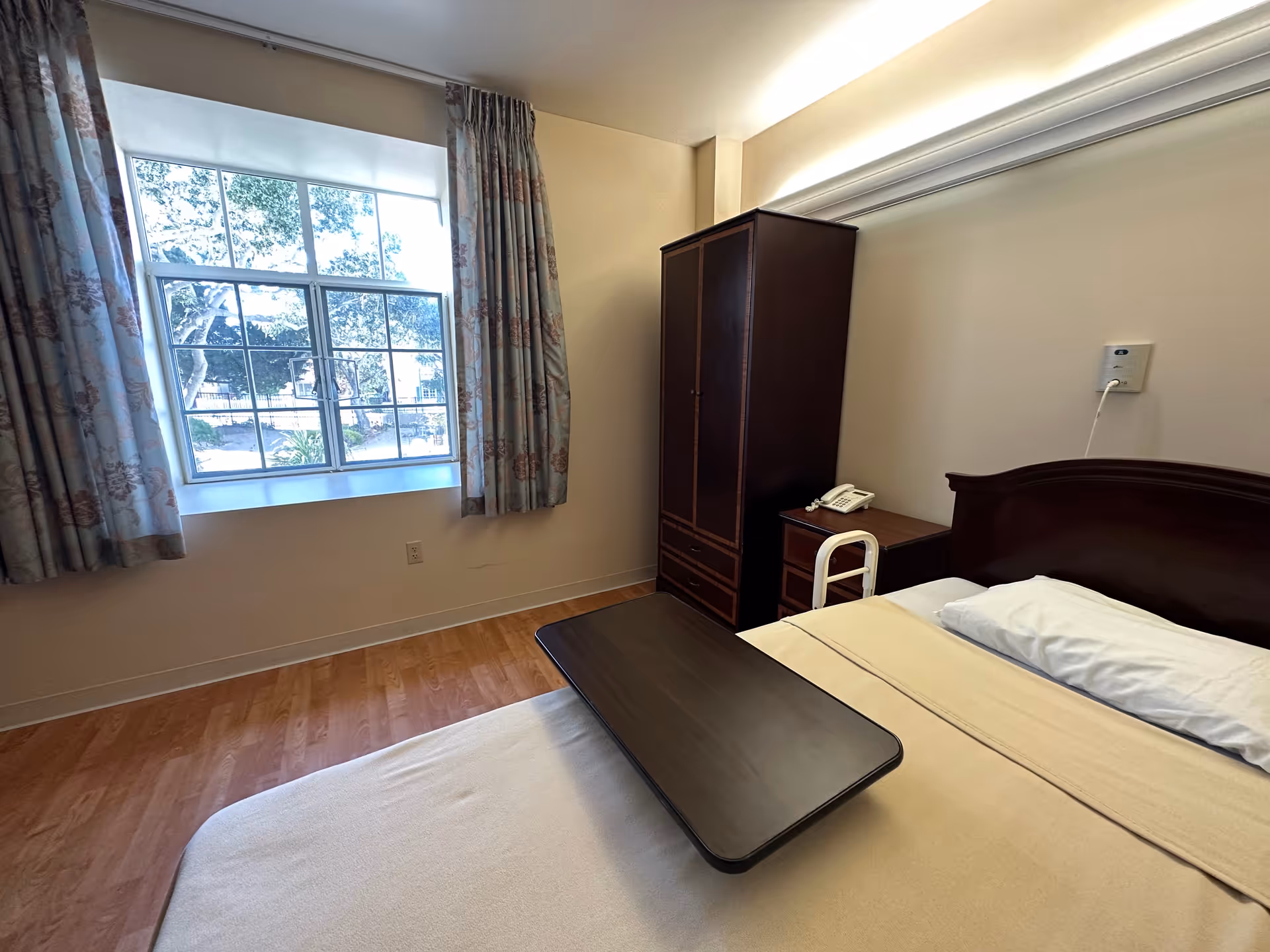 A senior bedroom with a bed, a small table, a wardrobe, and a window with curtains.
