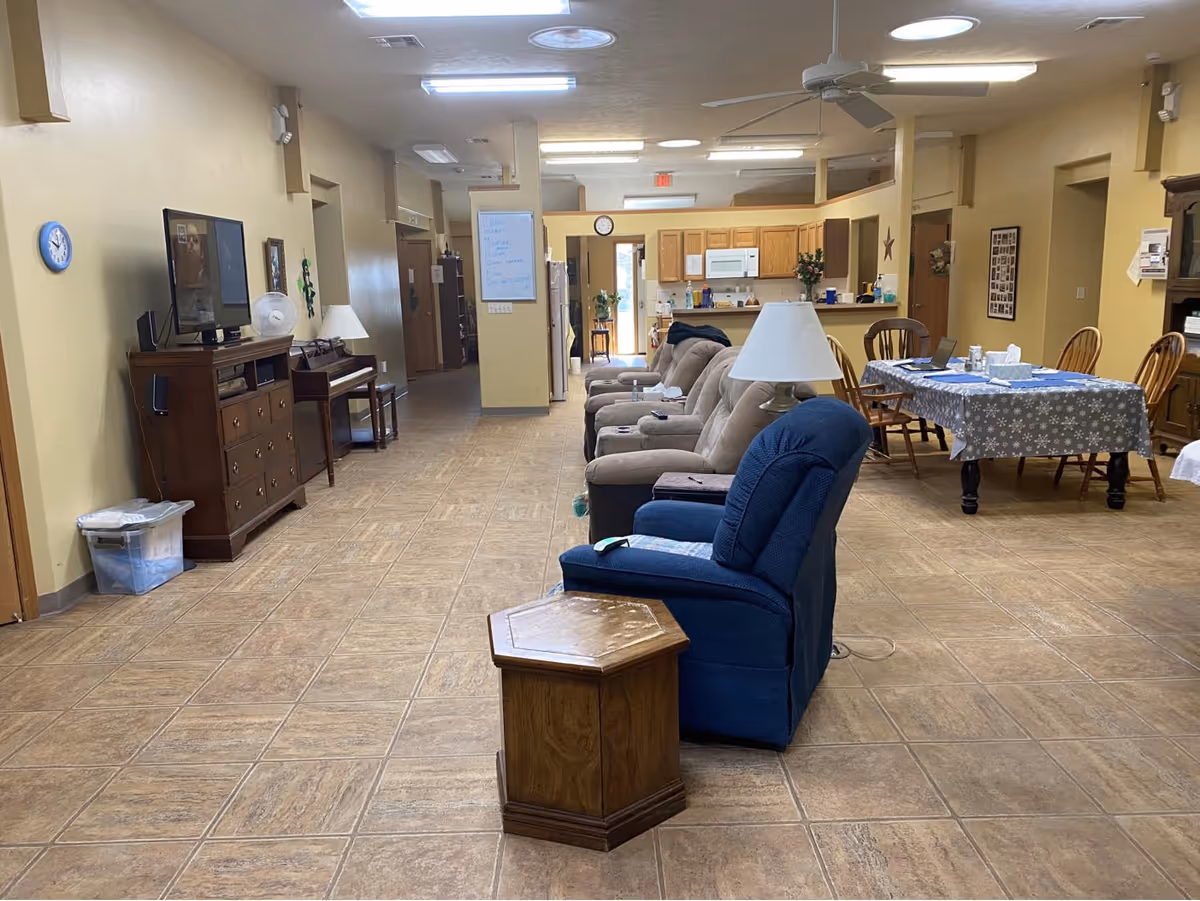 Interior view of a senior living facility common area with a row of recliner chairs and a blue armchair facing a TV on a wooden cabinet. There is a dining table with chairs covered with a tablecloth, a piano against the wall, and a kitchen area in the background. The floor is tiled and the walls are painted light yellow.
