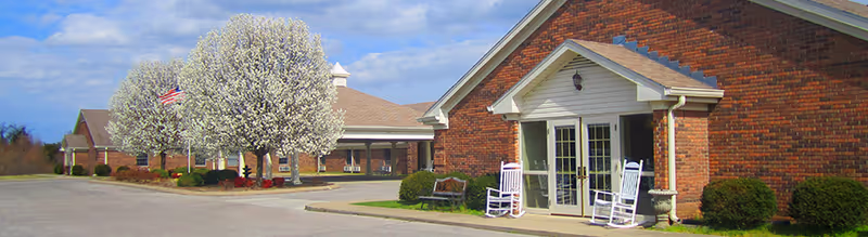 Exterior view of Village Manor Assisted Living facility showing a brick building with a covered entrance, two white rocking chairs on the porch, blooming white trees, an American flag, and a paved driveway.