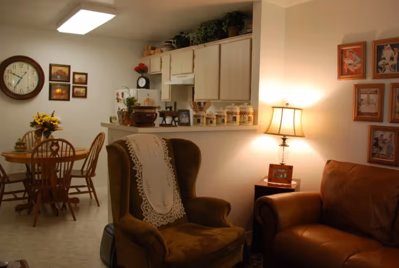 Cozy living room area with leather sofa, armchair, lamp, dining table, and open kitchen.