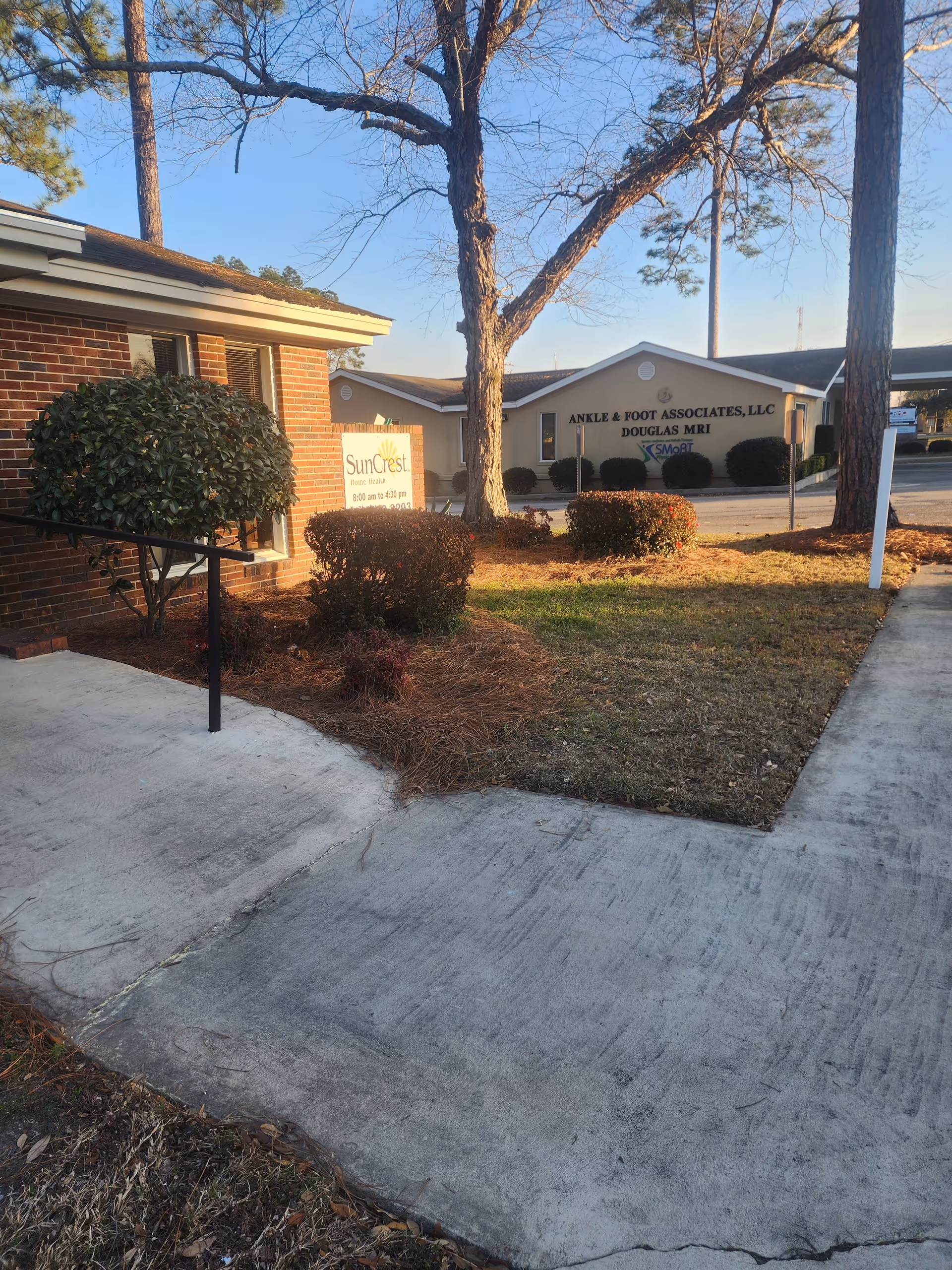 Exterior view of SunCrest Home Health facility with sign, shrubs, trees, and sidewalk.