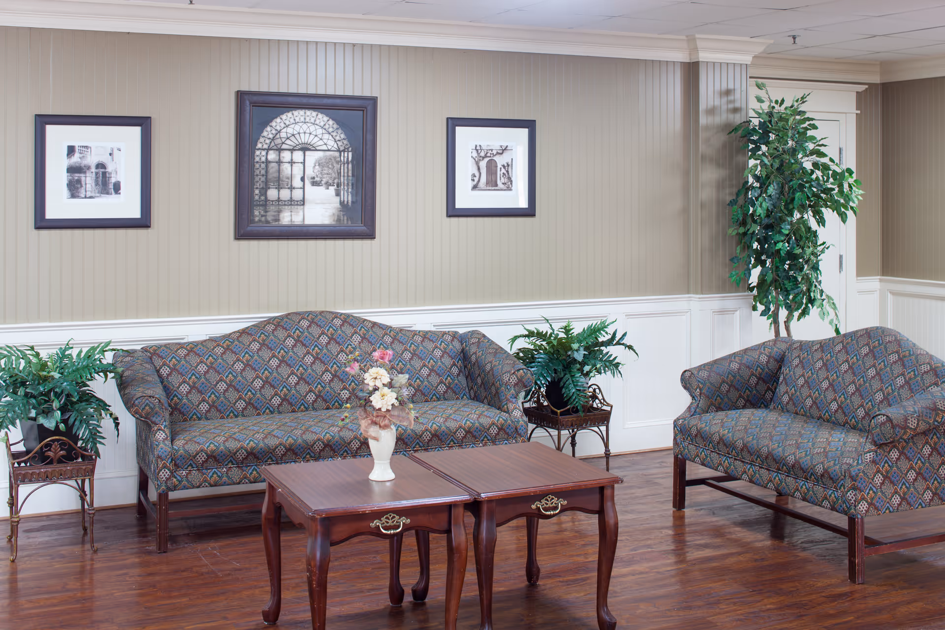 A sitting area with patterned sofas, a wooden coffee table, potted plants and framed artwork on a beige paneled wall.